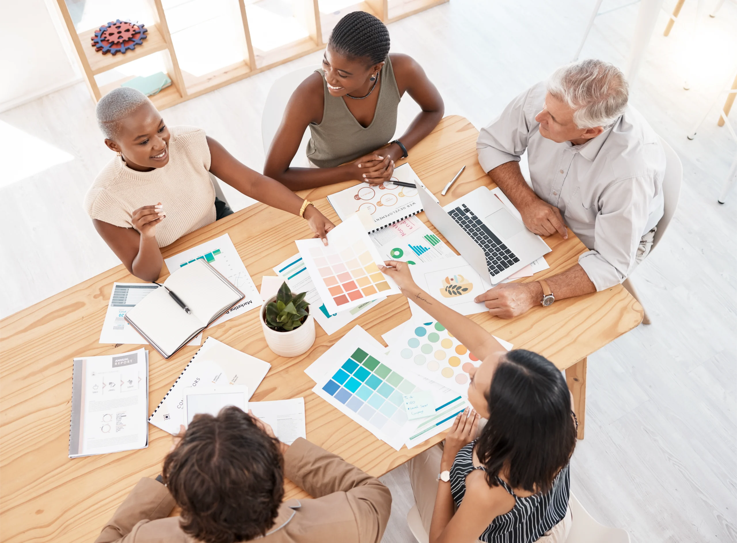 data-logistics-and-business-meeting-in-office-wit-2025-04-06-11-27-45-utc Men and women of different ages and backgrounds sitting at wooden table covered in printed out charts and other data. They are talking over the materials, including different color palettes.