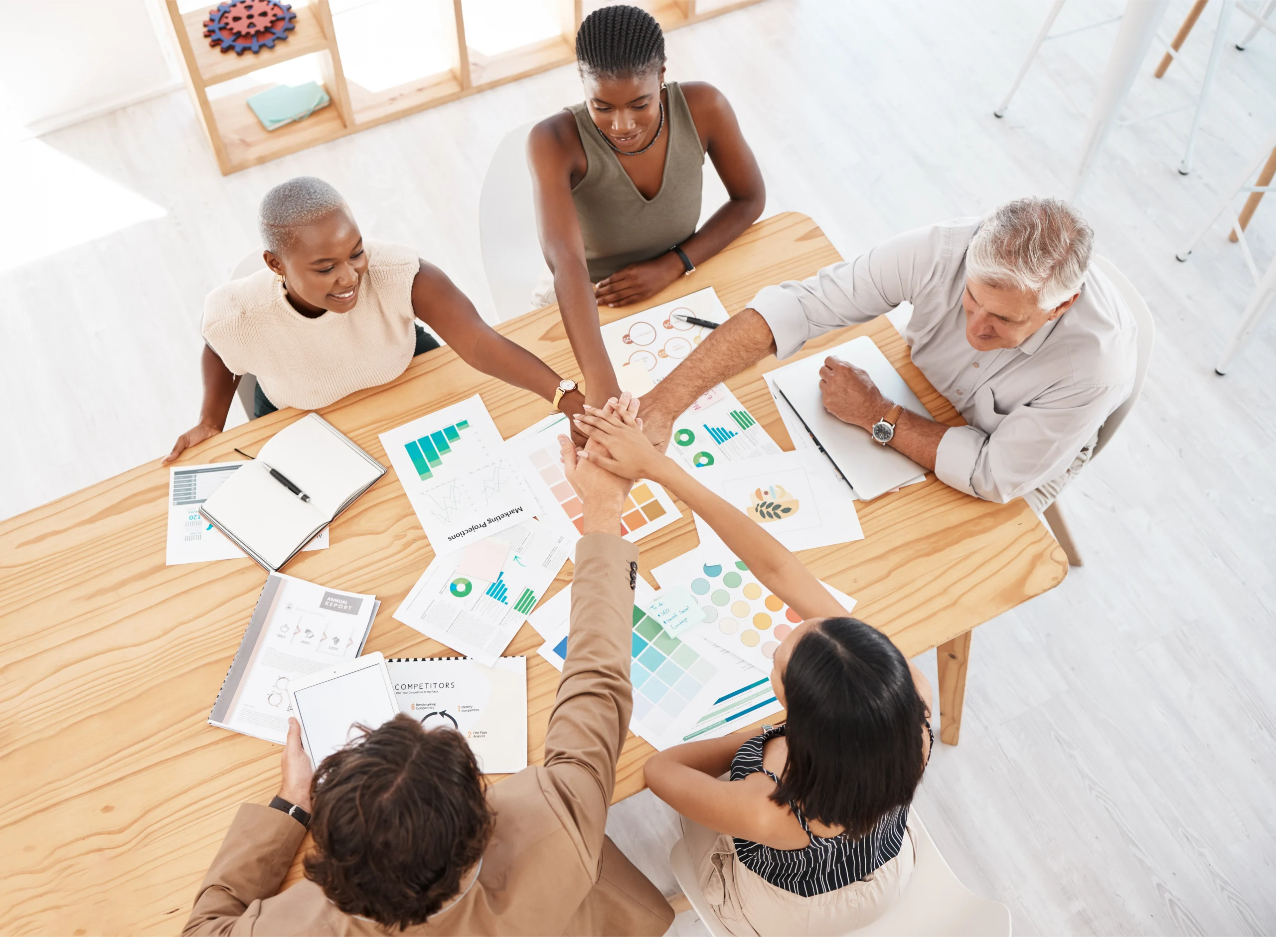 teamwork-meeting-and-hands-together-in-a-pile-aft-2025-04-06-07-49-39-utc Men and women of different ages and backgrounds sitting at wooden table covered in printed out charts and other data. Their hands are outstretched and meeting holding hands in the middle.